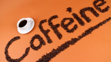 An image showing cup of black coffee along with coffee beans spelled into caffeine, a natural stimulant present in coffee, tea etc and is the cause of caffeine sensitivity (also called caffeine intolerance) in some people.